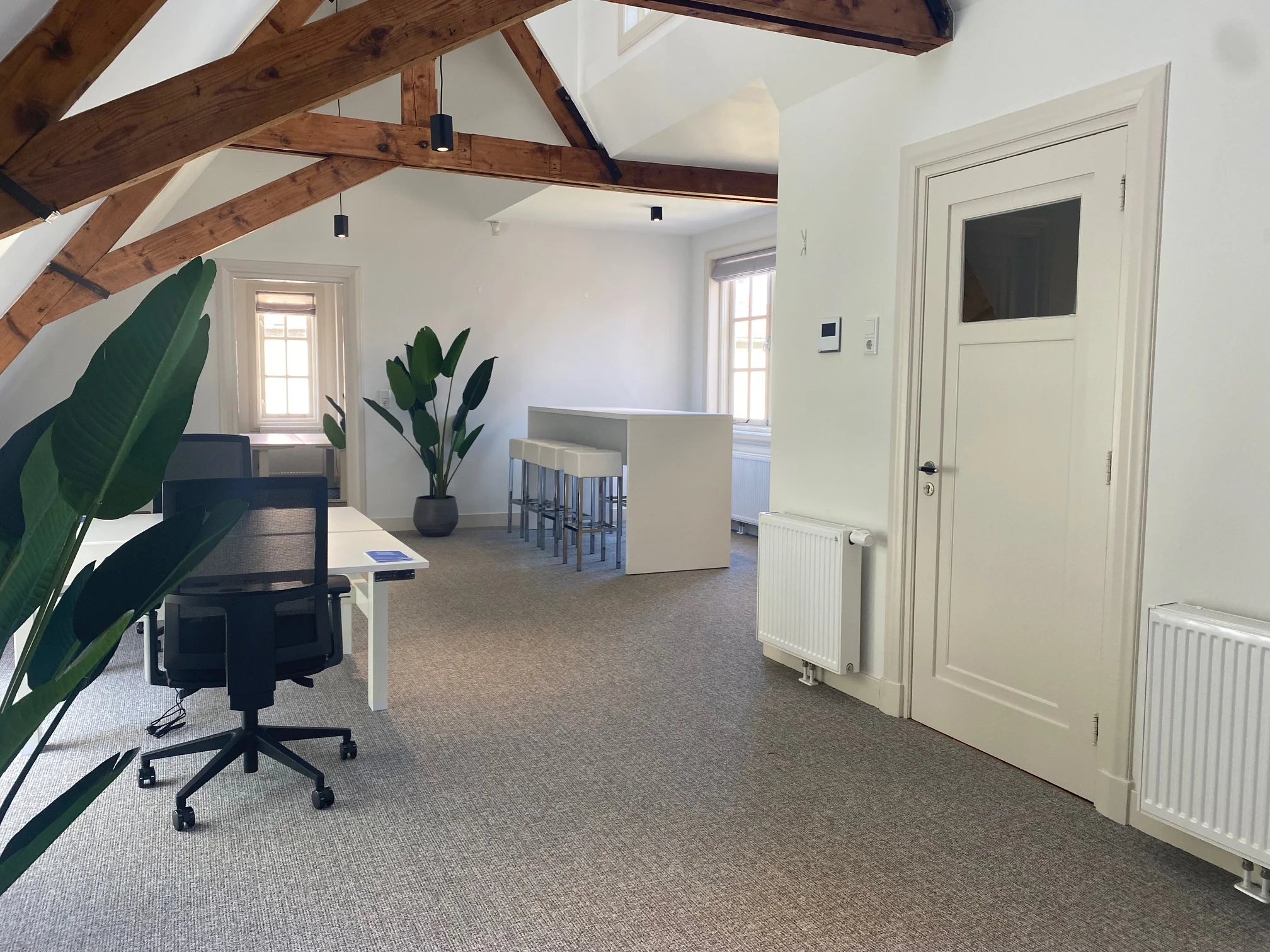 Modern kantoorinterieur aan de Kleine Houtweg met witte wanden, houten balken, bureaus, barkrukken en grote planten.